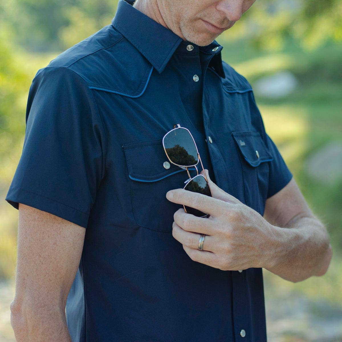 Man wearing a navy blue shirt holding sunglasses with a blurred green background