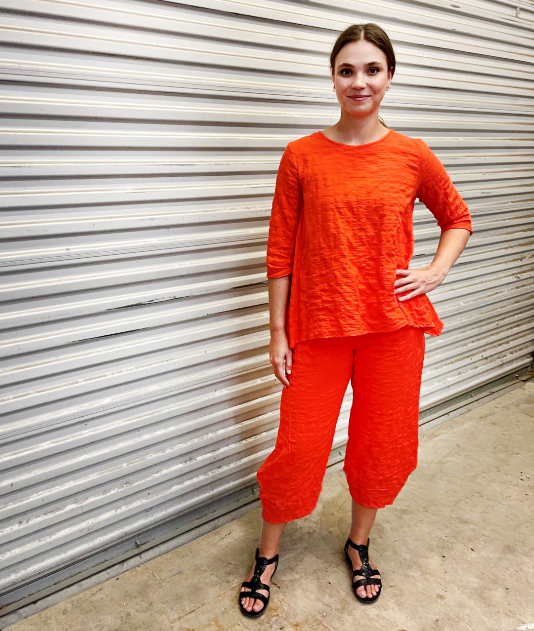 model in a chili red wide leg pant with a tapered ankle, worn with a matching pullover top with a 3/4 sleeve and dips in the hem at either hip