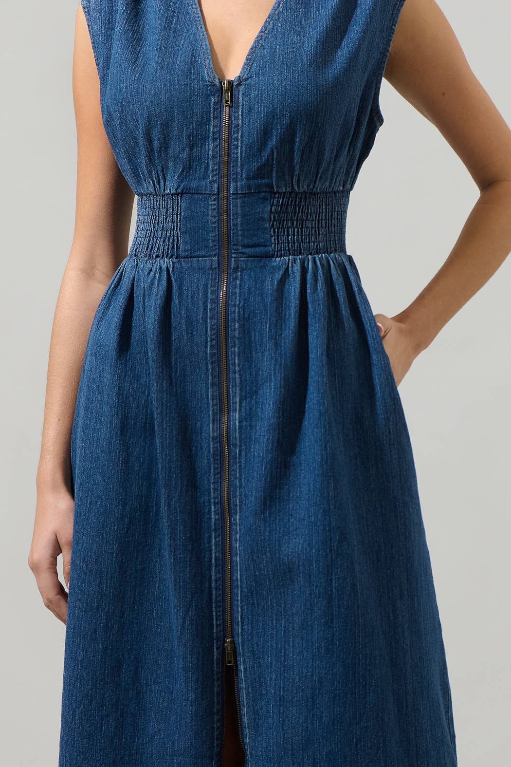 close up of model wearing a sleeveless midi denim dress with a v-neck, smocked waist and front zipper on a white background