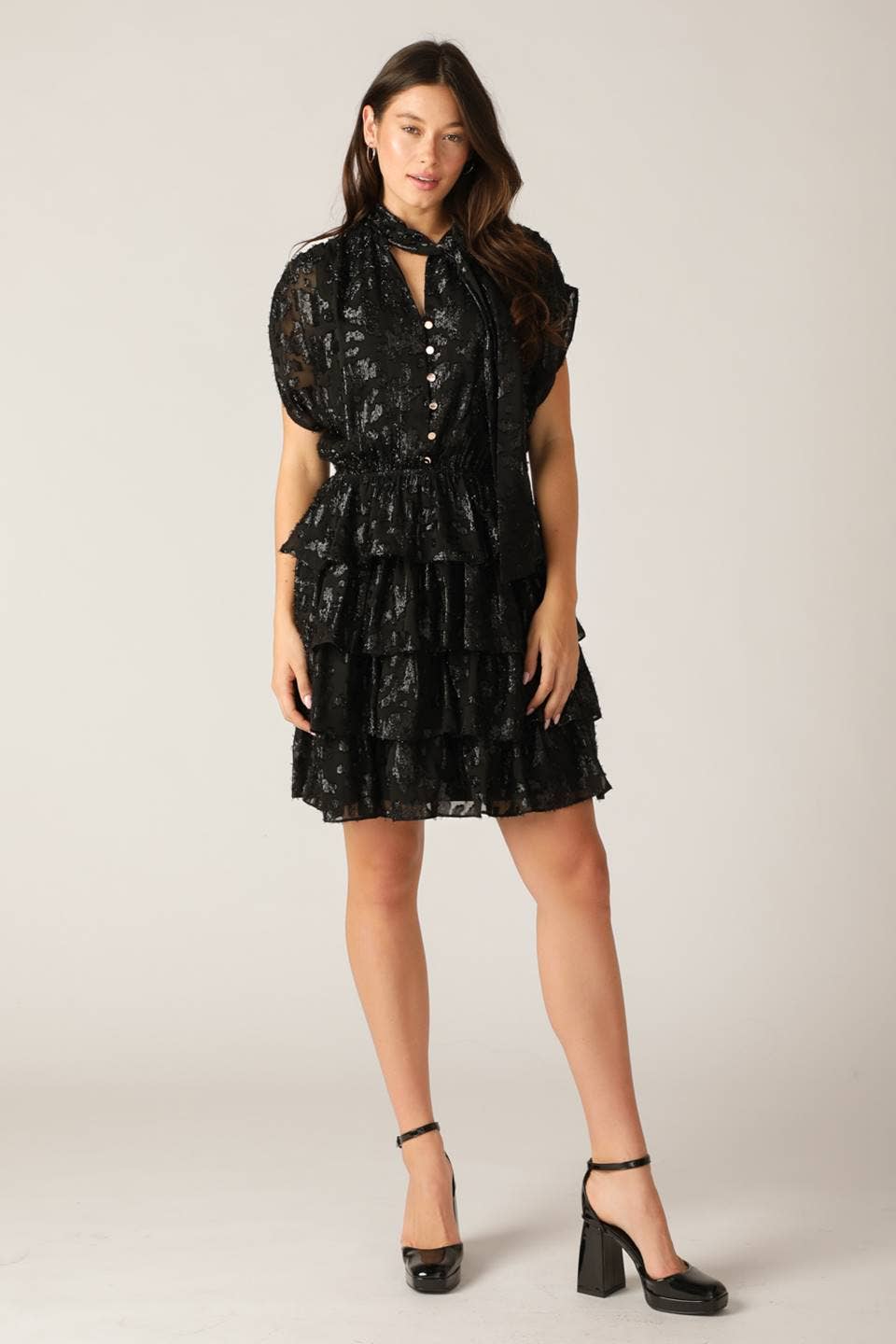model wearing a black shiny dress with a textured pattern featuring ruffle skirt and a neck tie on a white background