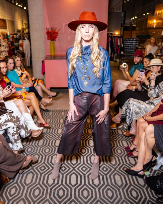model wearing orange hat, blue cowl top, statement necklace, brown shimmer stripe pants with taupe boots on runway