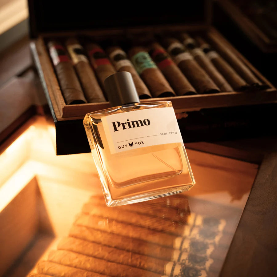 Perfume bottle labeled 'Primo' by Guy Fox on a wooden surface with a box of cigars in the background.