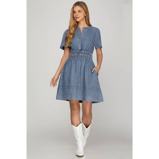 A woman wearing a blue washed denim short sleeve dress with elastic waistband and side pockets, paired with white boots.