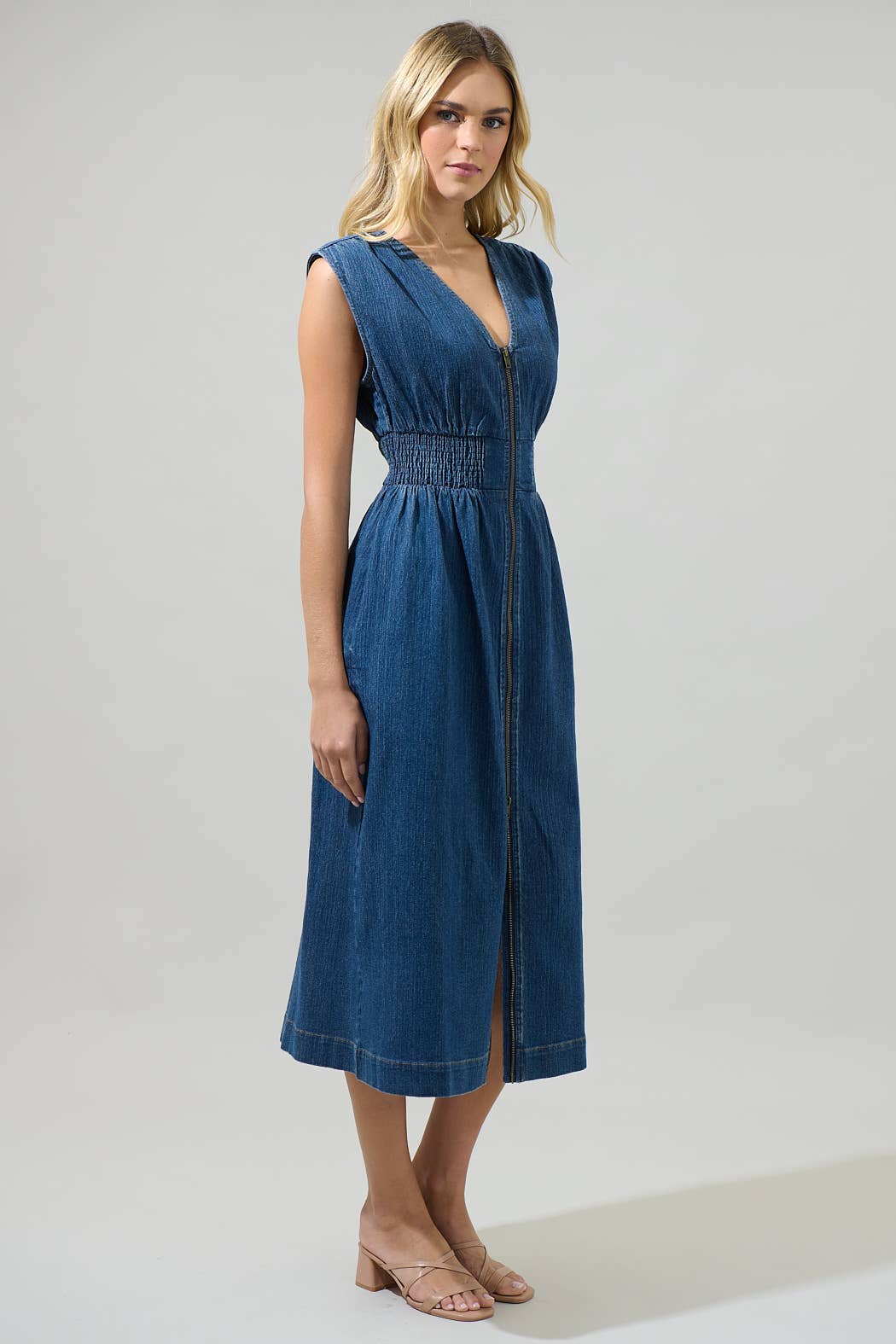 model wearing a sleeveless midi denim dress with a v-neck, smocked waist and front zipper on a white background