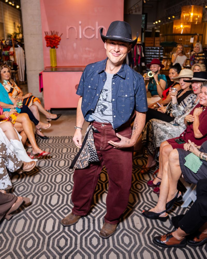 model wearing short sleeve navy pearl snap shirt with grey tshirt underneath, burgundy pants, black cowboy hat and brown cowboy boots on a runway