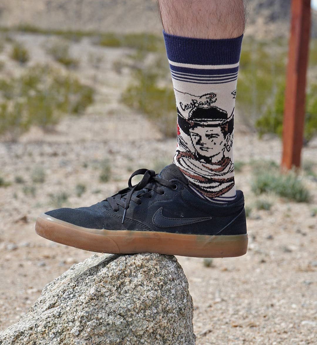 mens sock with navy and red stripe and black and white cowboy drawing. black sneaker and a rustic outdoor background