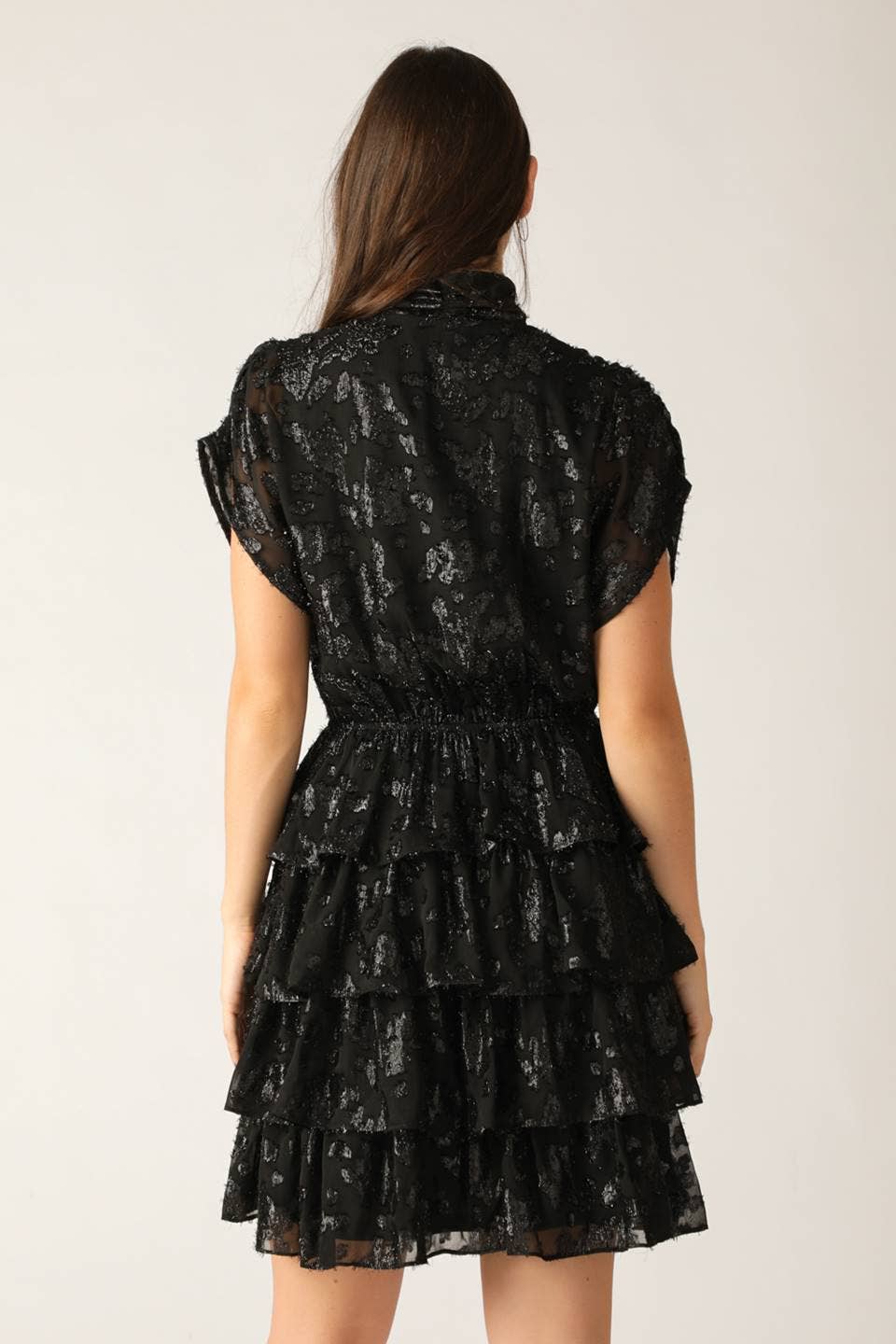 model showing back of a black shiny dress with a textured pattern featuring ruffle skirt and a neck tie on a white background