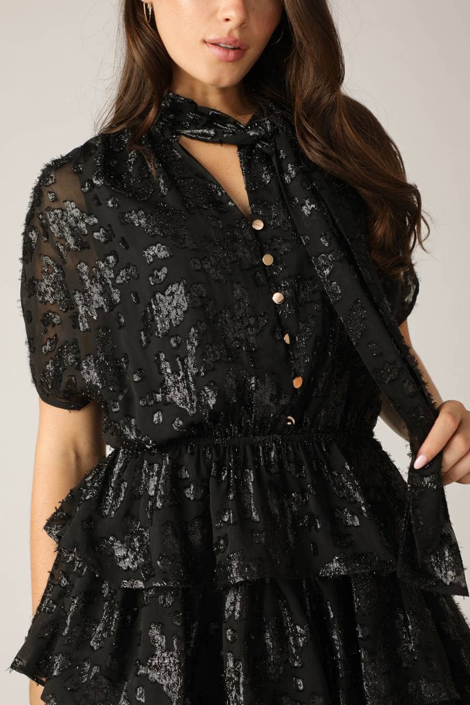 close up of model wearing a black shiny dress with a textured pattern featuring ruffle skirt and a neck tie on a white background