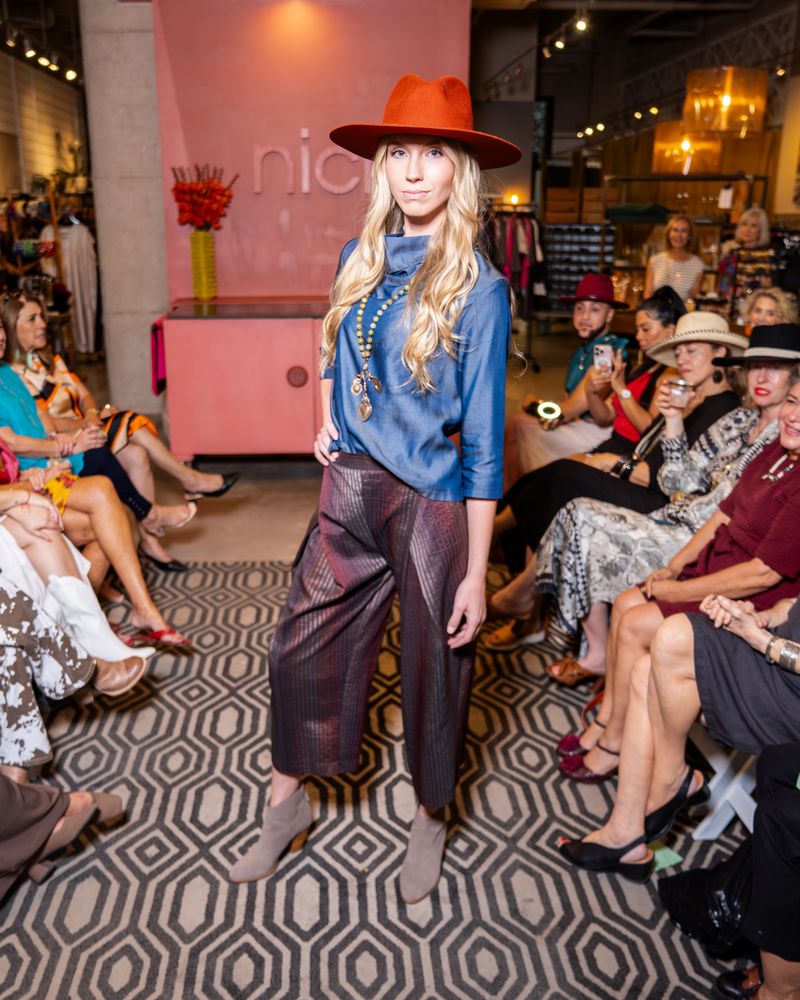 model wearing orange hat, blue cowl top, statement necklace, brown shimmer stripe pants with taupe boots on runway
