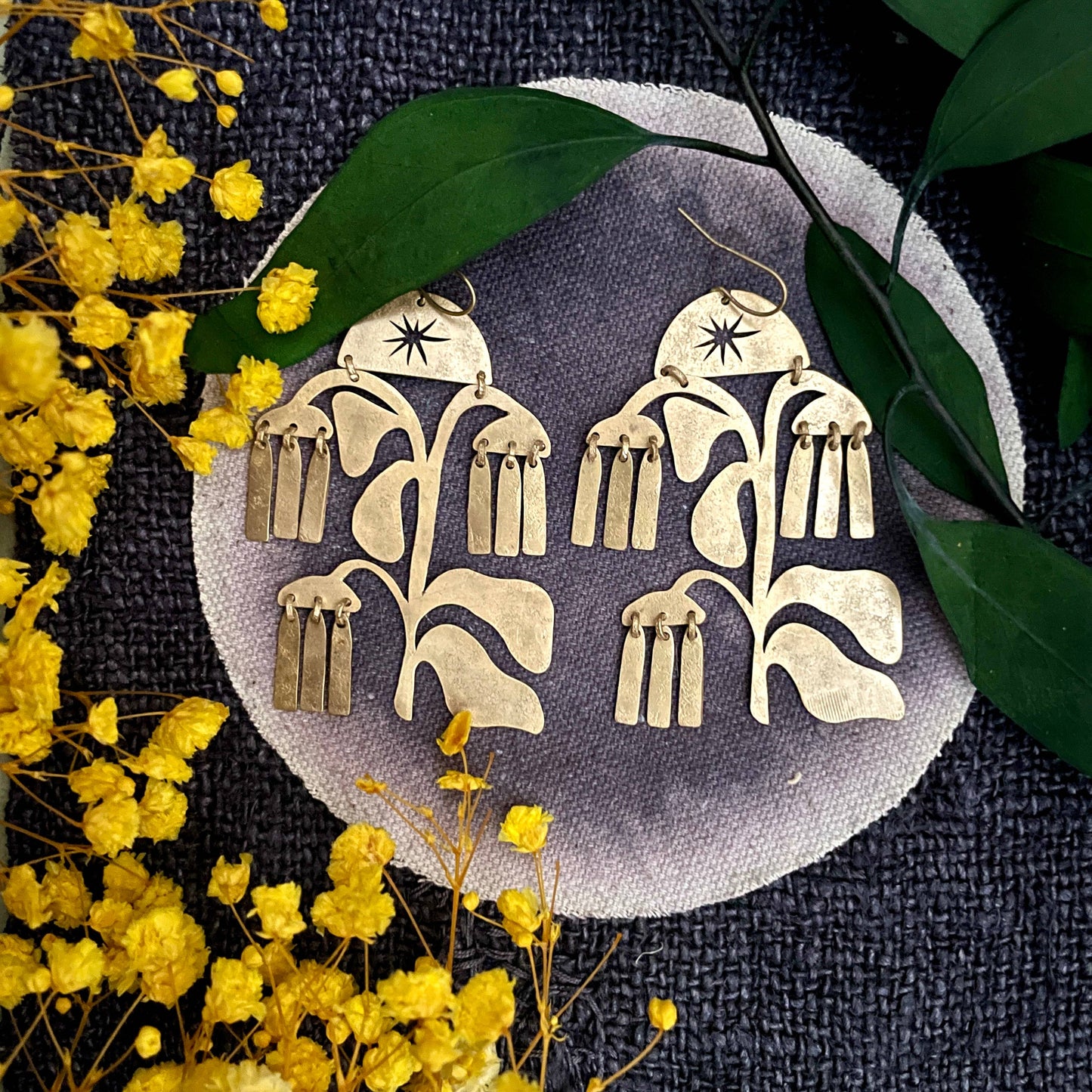 Gold floral earrings on a dark fabric background with yellow flowers and green leaves.