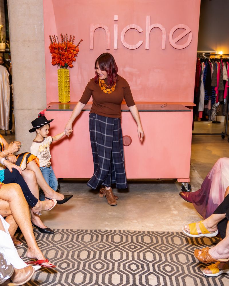 woman wearing brown knit top with layered necklace, navy plaid pants and boots holding hand of little boy wearing black cowboy hat, "yeehaw" shirt and jeans on a runway
