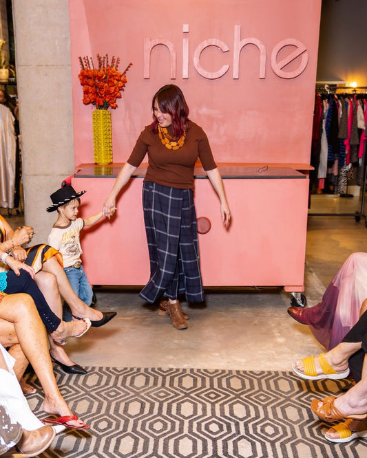 woman wearing brown knit top with layered necklace, navy plaid pants and boots holding hand of little boy wearing black cowboy hat, "yeehaw" shirt and jeans on a runway