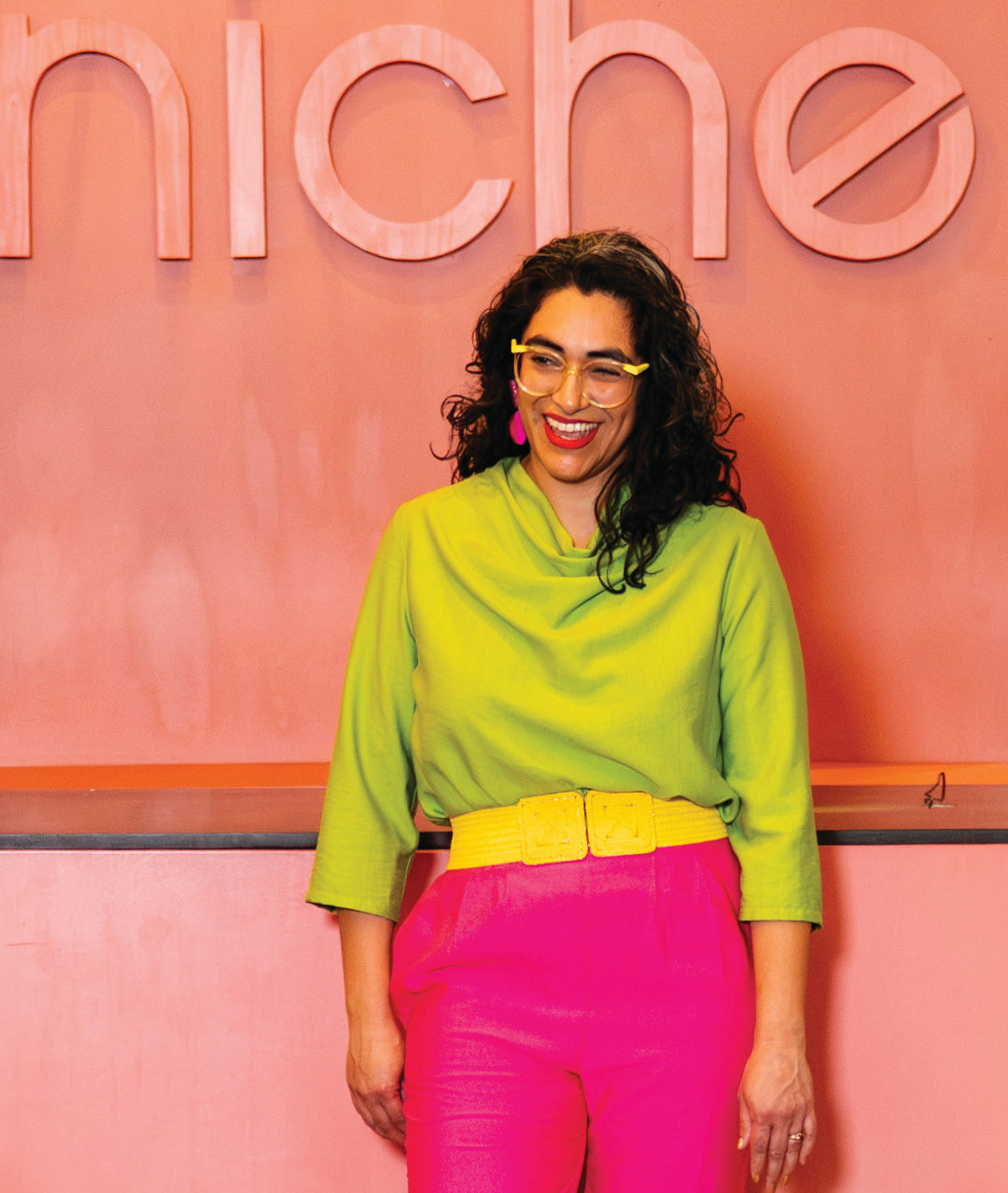 model in a lime top with a 3/4 sleeve and cowl neck, worn with a hot pink crop pant and a wide yellow belt