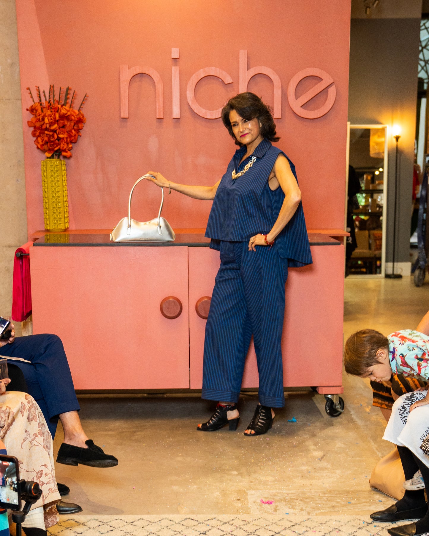 Woman in a blue top and pant and silver necklace and purse walking down a runway at a fashion event