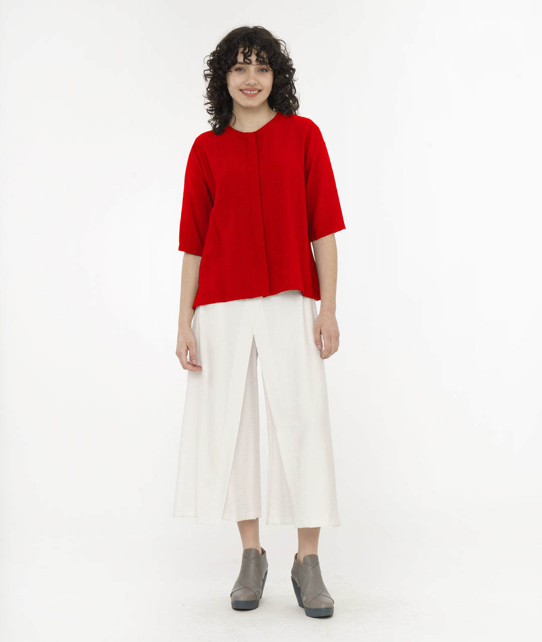 model in a red top with a hidden button placket. top has a round neckline and a 3/4 sleeve, with a small split at each hip side seam. worn with a white wide leg pant with overlapping panels
