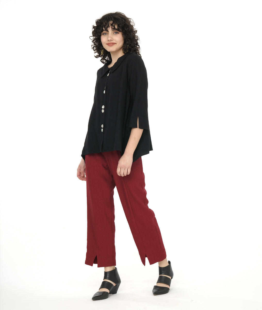 model in a black button down jacket with a 3/4 sleeve, dipped hem at the sides, and a twin button detail at the back and placket. worn with a straight leg deep red pant with a split at each ankle
