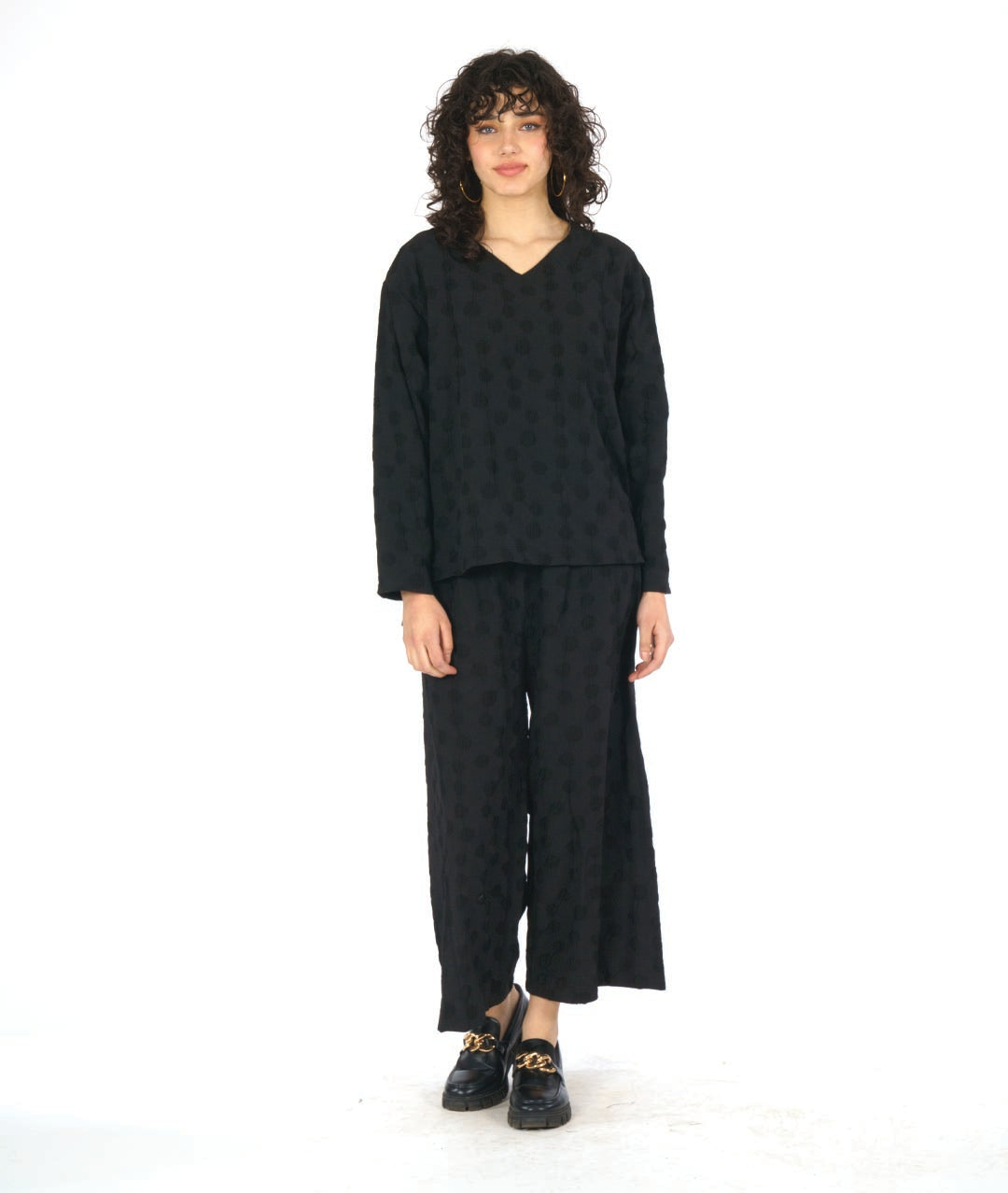 model in a black pullover blouse with a puckered dot texture. top has long sleeves, a v-neck and a stairstep hem. worn with a wide leg matching pant