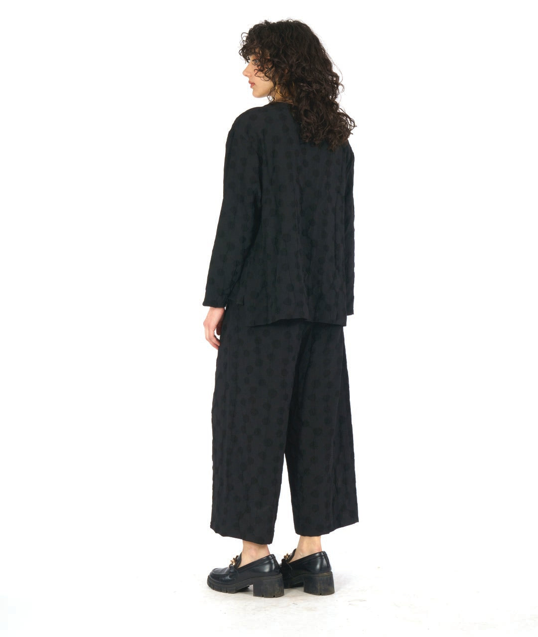 model in a black pullover blouse with a puckered dot texture. top has long sleeves, a v-neck and a stairstep hem. worn with a wide leg matching pant