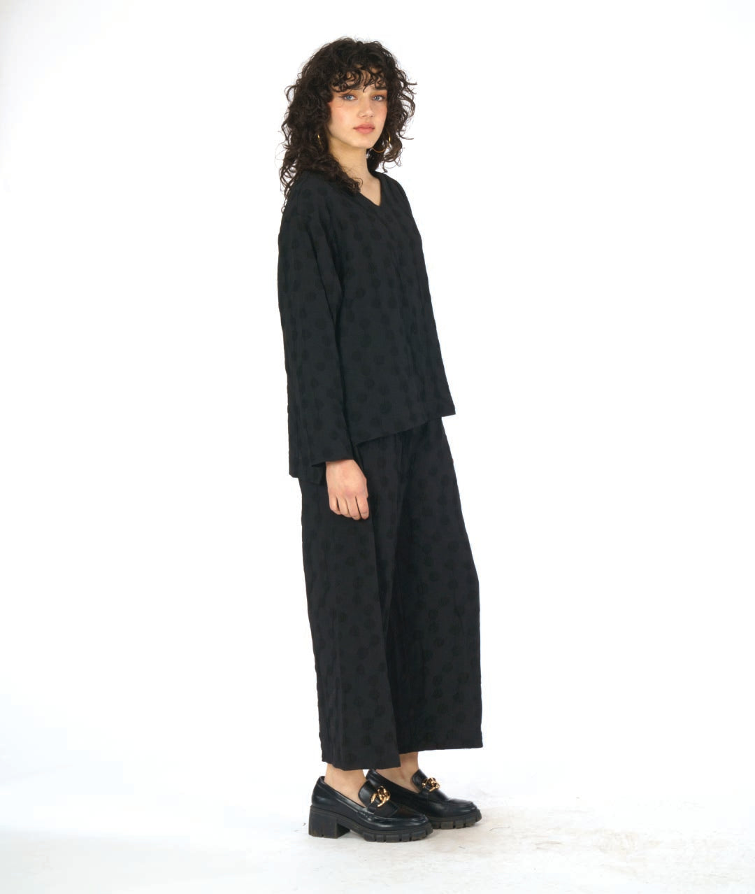 model in a black pullover blouse with a puckered dot texture. top has long sleeves, a v-neck and a stairstep hem. worn with a wide leg matching pant