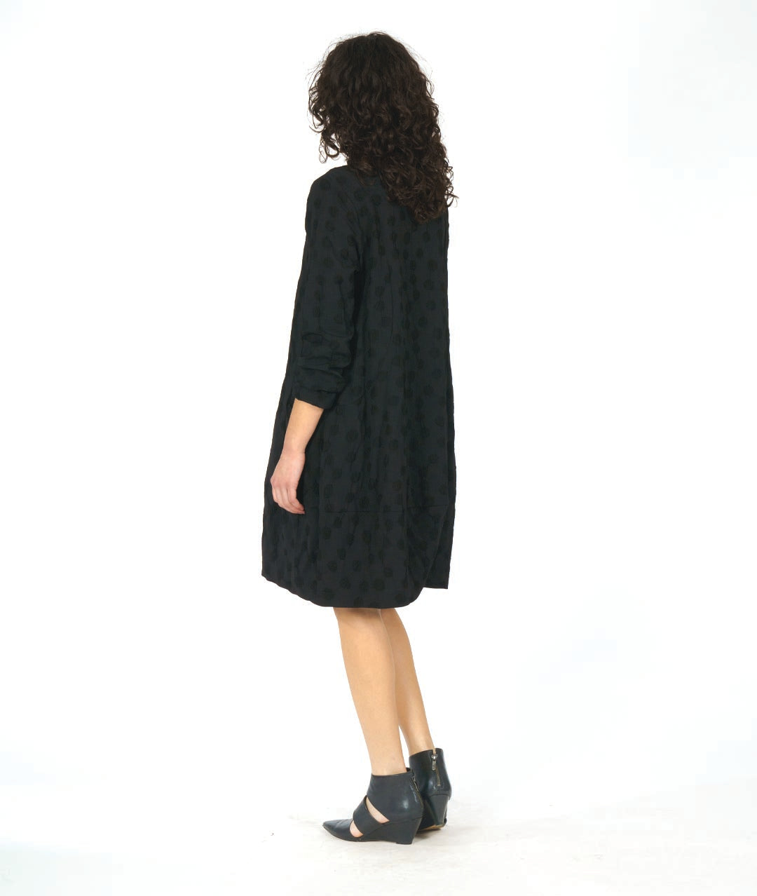 model in a black pullover, knee length dress with long sleeves and circular seams at the hips with a pocket