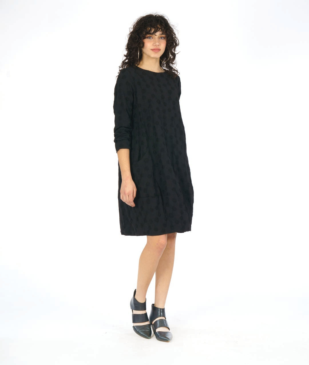 model in a black pullover, knee length dress with long sleeves and circular seams at the hips with a pocket