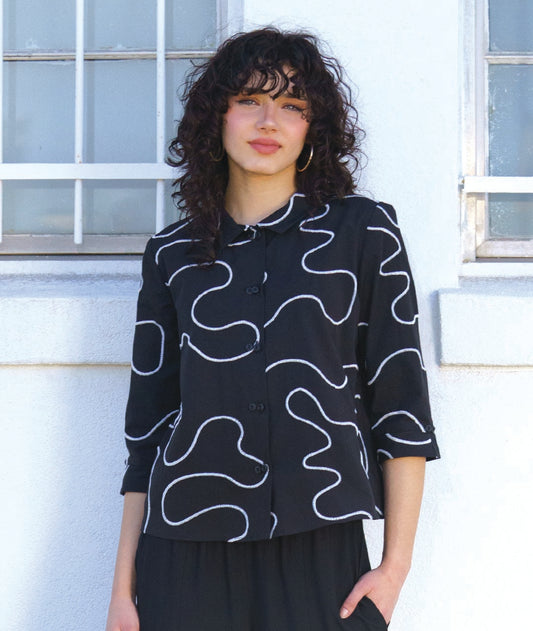 model in a black pant with a black button down with a white swirl embroidery and a twin button detail