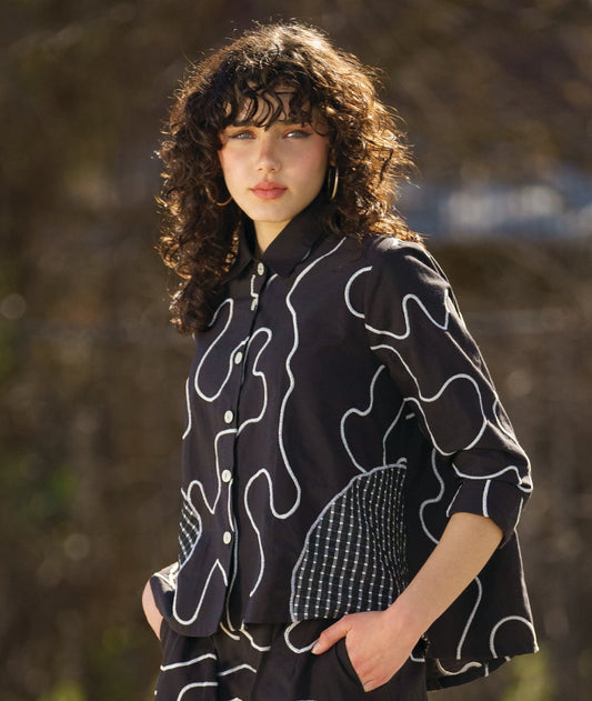 model wearing a black button down blouse with a bell shaped wide body. blouse has a white swirl print embroidered all over, and large round patches of check print fabric accented across