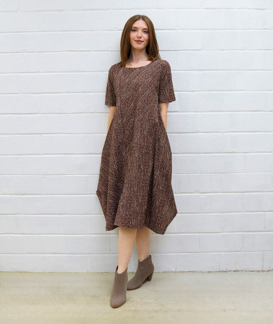 Woman wearing a brown patterned dress standing against a white brick wall.