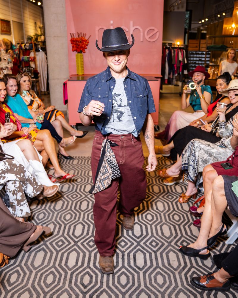 model wearing short sleeve navy pearl snap shirt with grey tshirt underneath, burgundy pants, black cowboy hat and brown cowboy boots on a runway