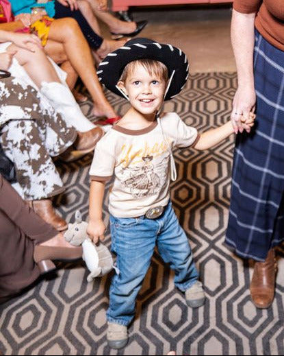 woman wearing brown knit top with layered necklace, navy plaid pants and boots holding hand of little boy wearing black cowboy hat, "yeehaw" shirt and jeans on a runway