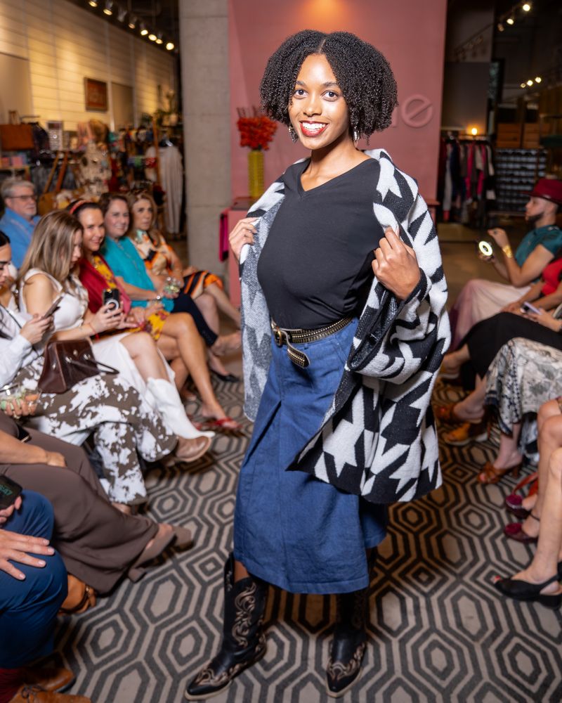 model wearing black and white houndstooth cardigan, black top, belt, jean skirt and cowboy boots on a runway