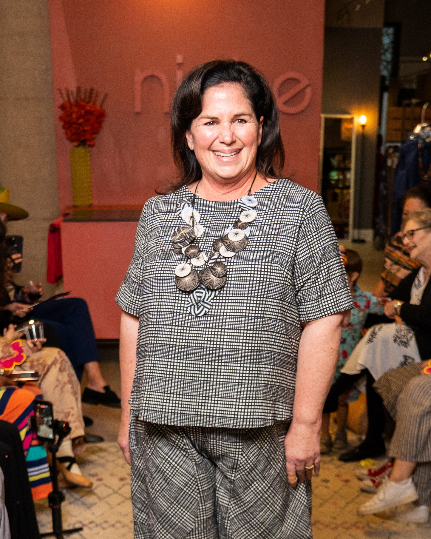 Woman in a checkered outfit with a necklace standing in a social setting.