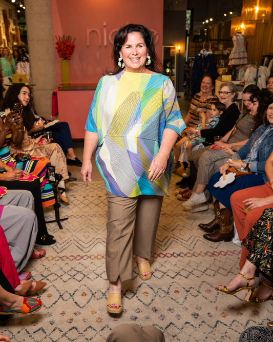 Woman in a colorful top and taupe pants walking down a runway.
