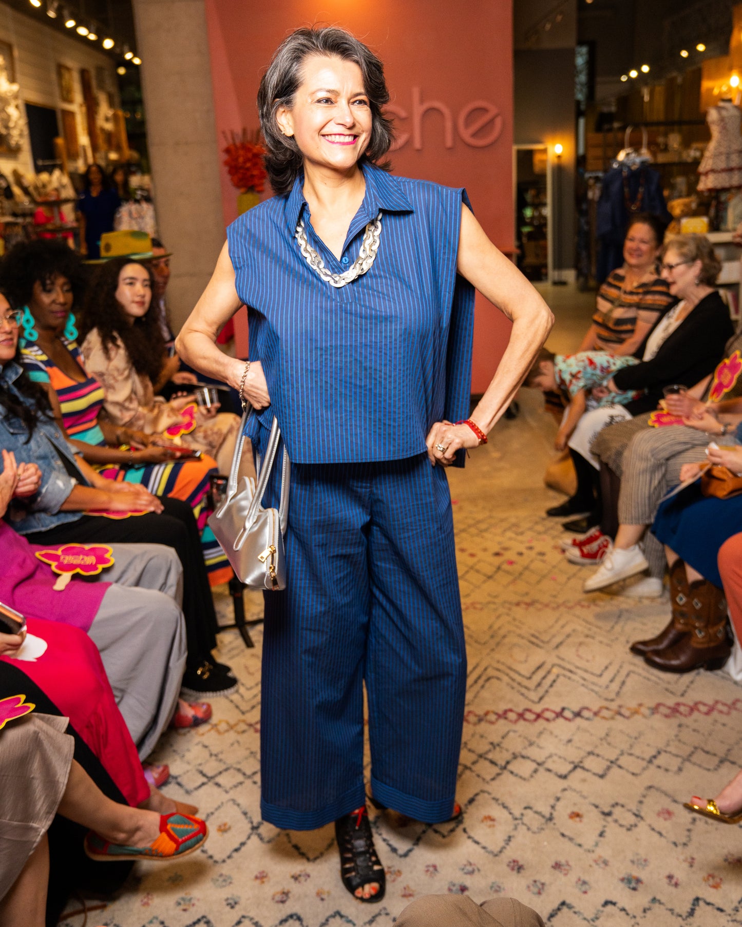 Woman in a blue top and pant and silver necklace and purse walking down a runway at a fashion event