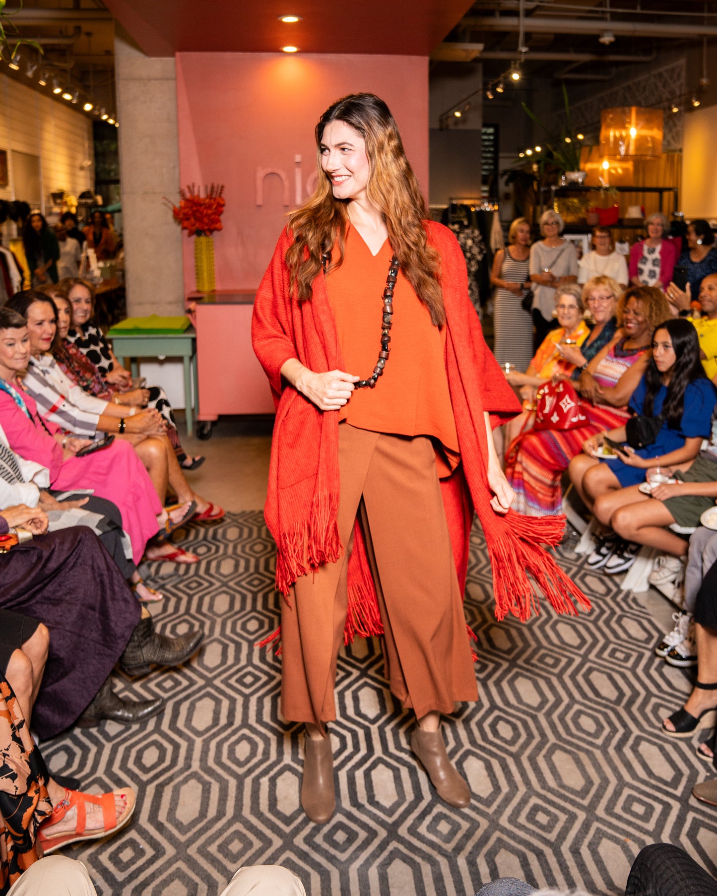 model walking down runway wearing orange v-neck top with wide leg nutmeg colored pants that have an overlapping layer in the front, burnt red cardigan and brown shoes