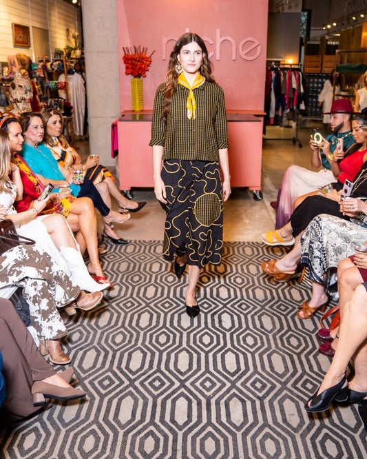 Model walking on a catwalk at a fashion event with wearing yellow check button up top, swirl pattern pants and yellow bandana