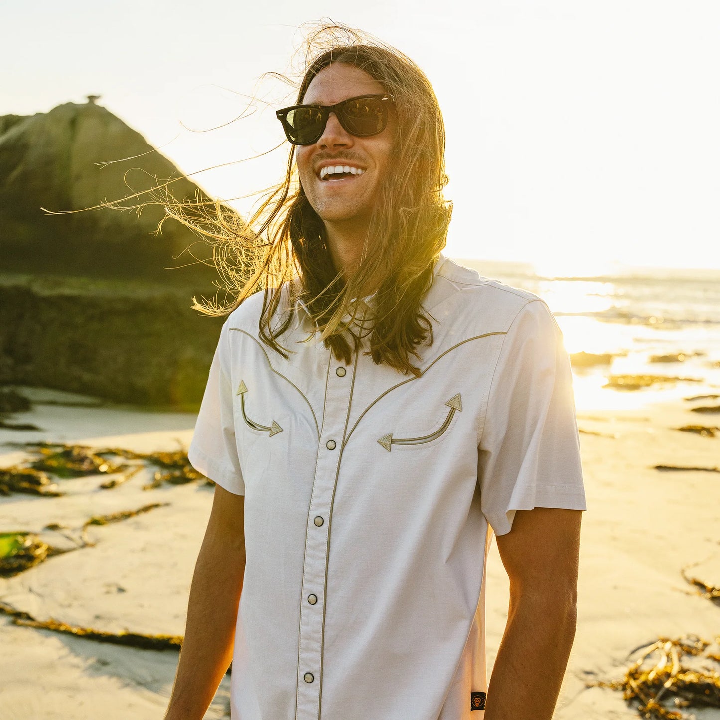 Man with sunglasses and long hair wearing a western short-sleeve shirt standing on a beach