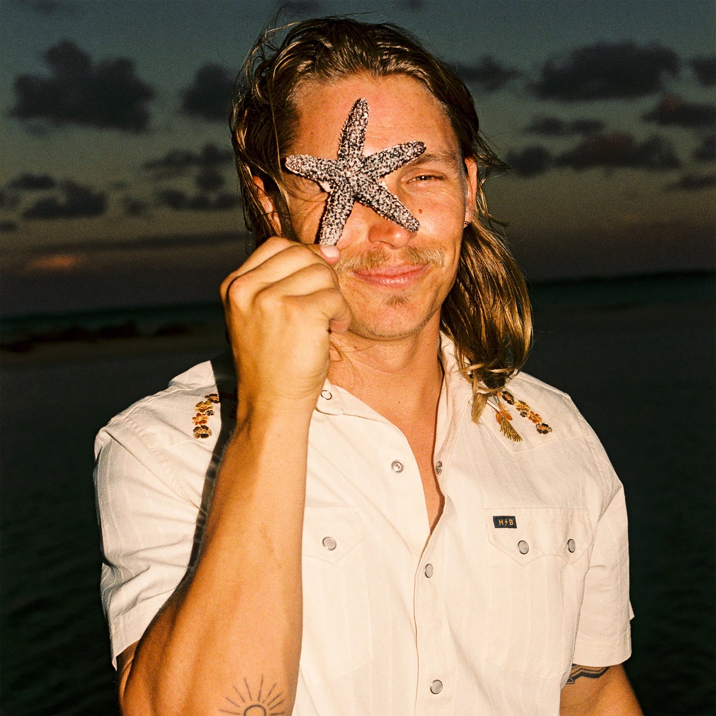 Man holding a starfish over his eye and wearing a beige short-sleeve button up shirt with a sunset or sunrise background