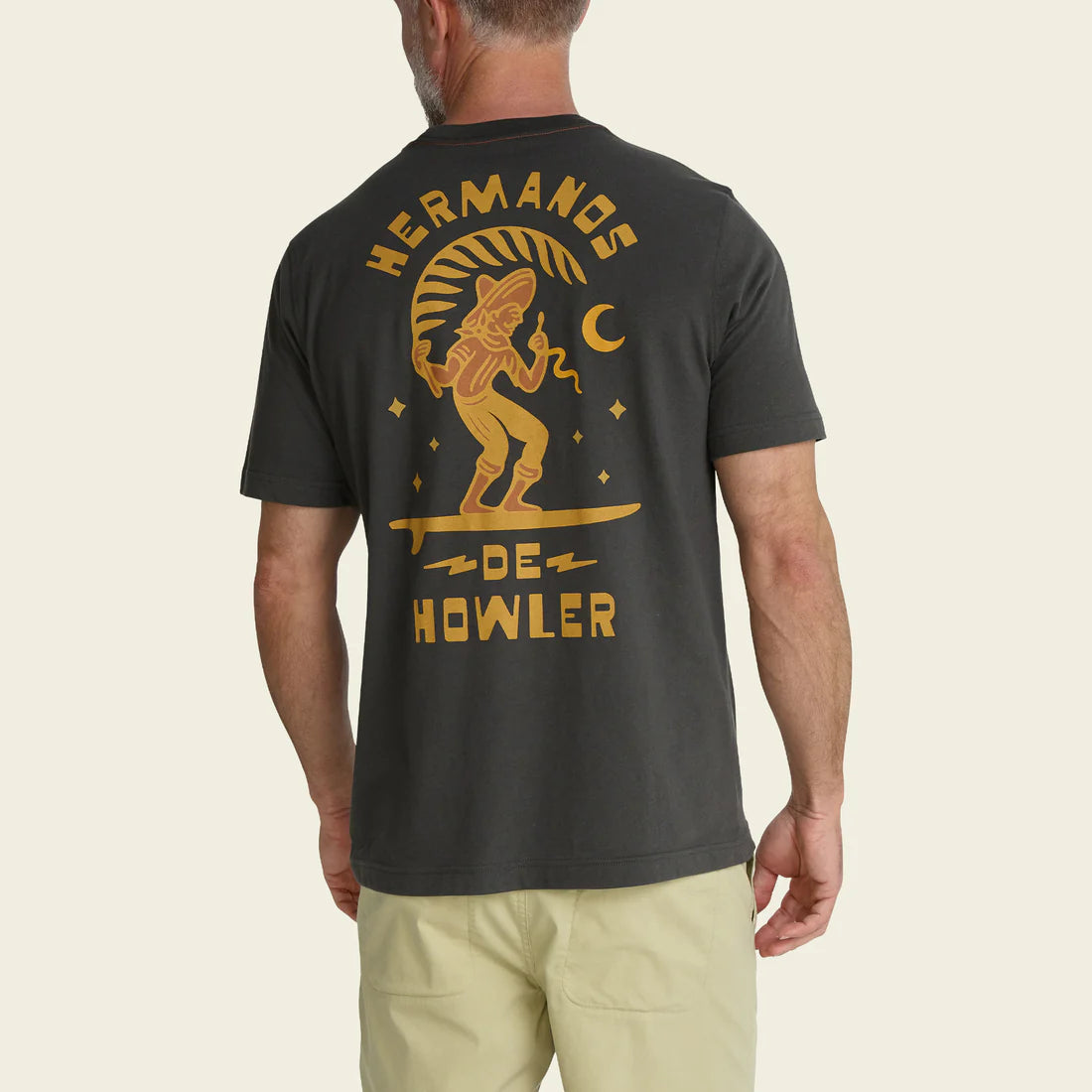 Man wearing a black t-shirt with gold graphic and text saying "HERMANOS DE HOWLER" on the back, standing against a beige background
