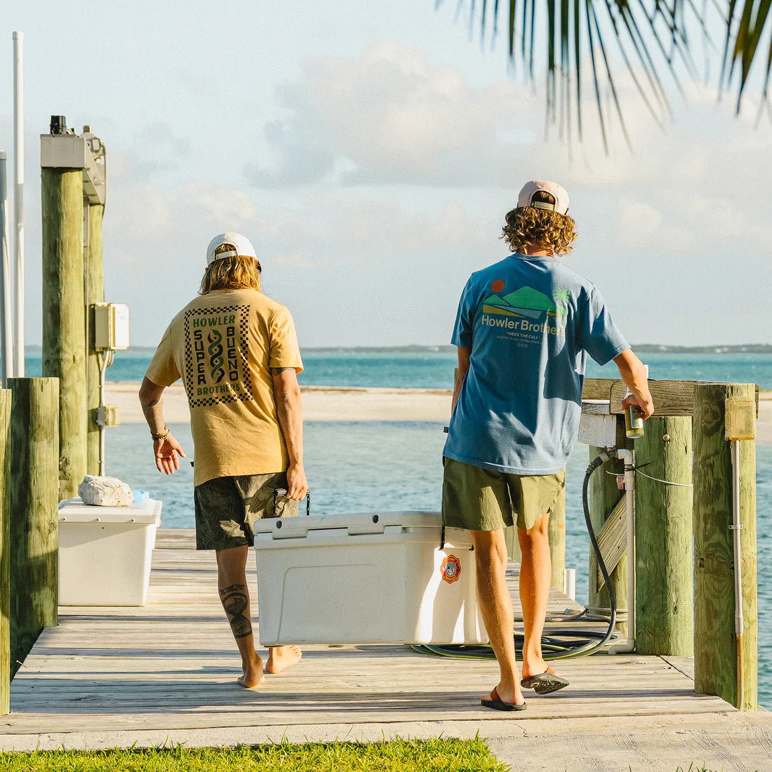 Two people carrying a cooler on a dock by the water wearing howler brother tshirts, hats and shorts