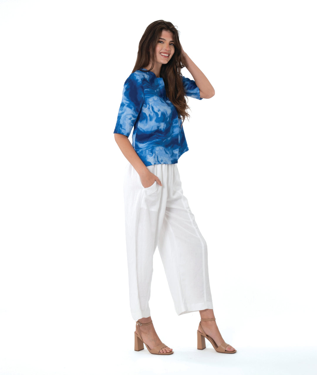 A woman modeling a blue and white abstract patterned short-sleeve top paired with white wide-leg cropped pants and tan heeled sandals, photographed against a seamless white studio background.