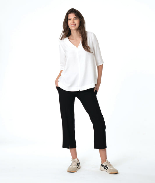 A woman modeling a white V-neck blouse with three-quarter sleeves and black cropped pants, paired with beige sneakers, against a solid white studio background.