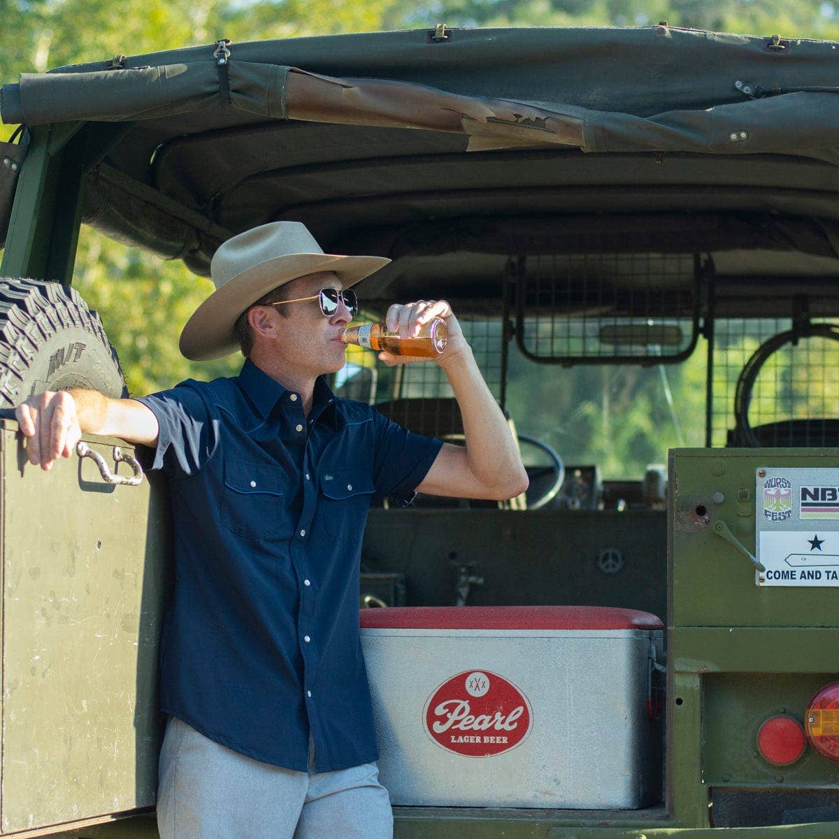 Man in cowboy hat drinking from a beer mug next to a green vehicle with a Pearl cooler.
