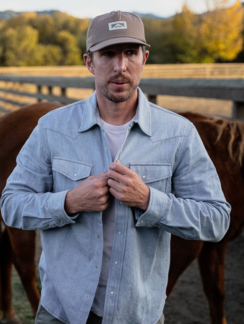 Man wearing a light blue shirt and brown cap standing in front of horses in a field.