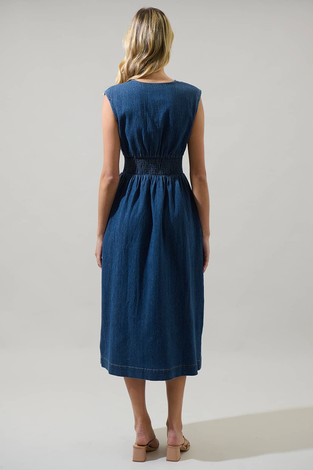 model showing back of sleeveless midi denim dress with smocked waist on a white background