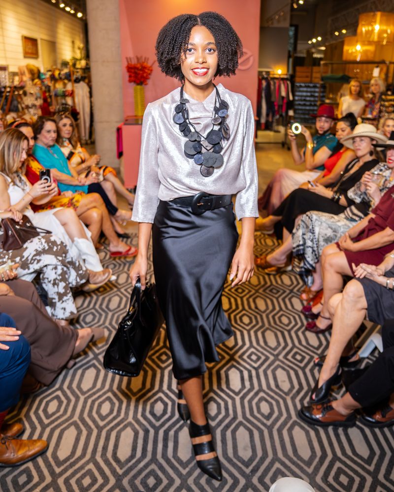 model wearing a silver shimmery blouse, platillos necklace, belt, purse and black satin skirt walking down a runway