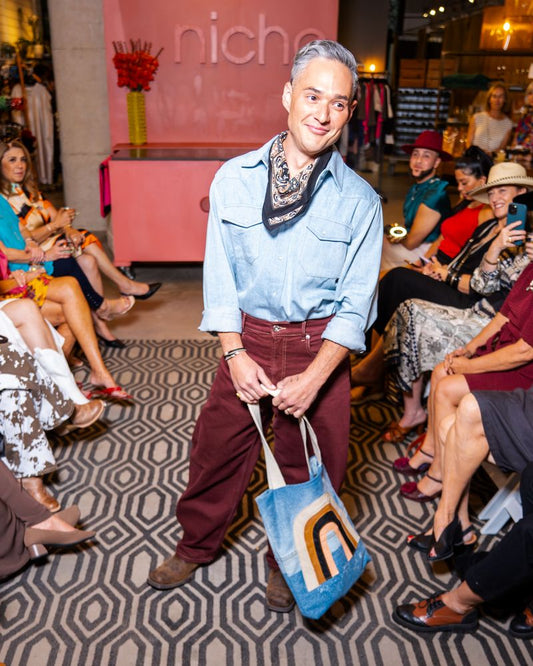 model wearing light blue pearl snap shirt with bandana, burgundy pants and jean tote with rainbow design on a runway