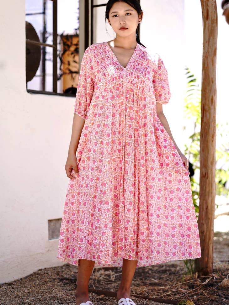 Close up of a front facing model wearing a v-necked pink and ivory printed dress w/ elbow length sleeves and tuck detail from shoulder to bust. Background outdoor of a white house