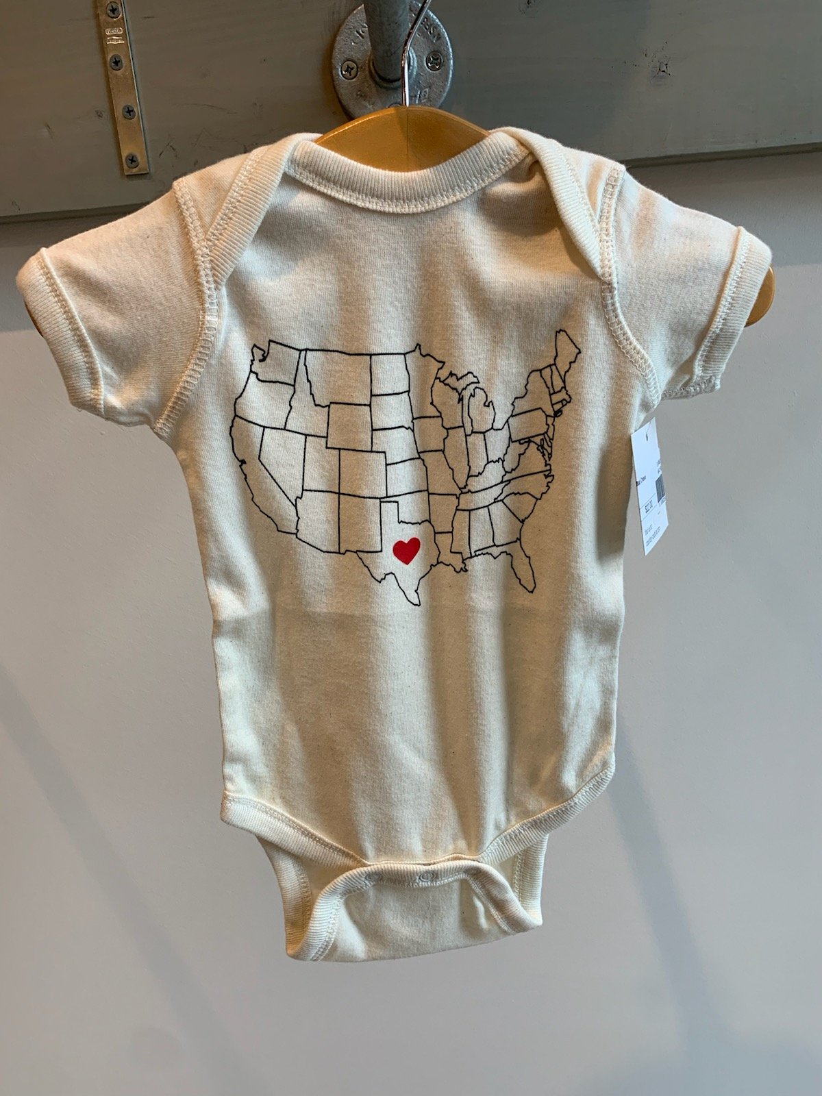 an ivory baby onesie with a drawing of  the united states with a heart in the center of texas