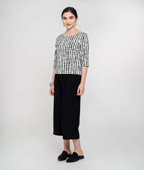 Pictured against a white background is a light skinned model with dark hair is smiling and wearing a 3/4 sleeved black and white dot jersey top and midi-length black pants and black slip-on shoes.